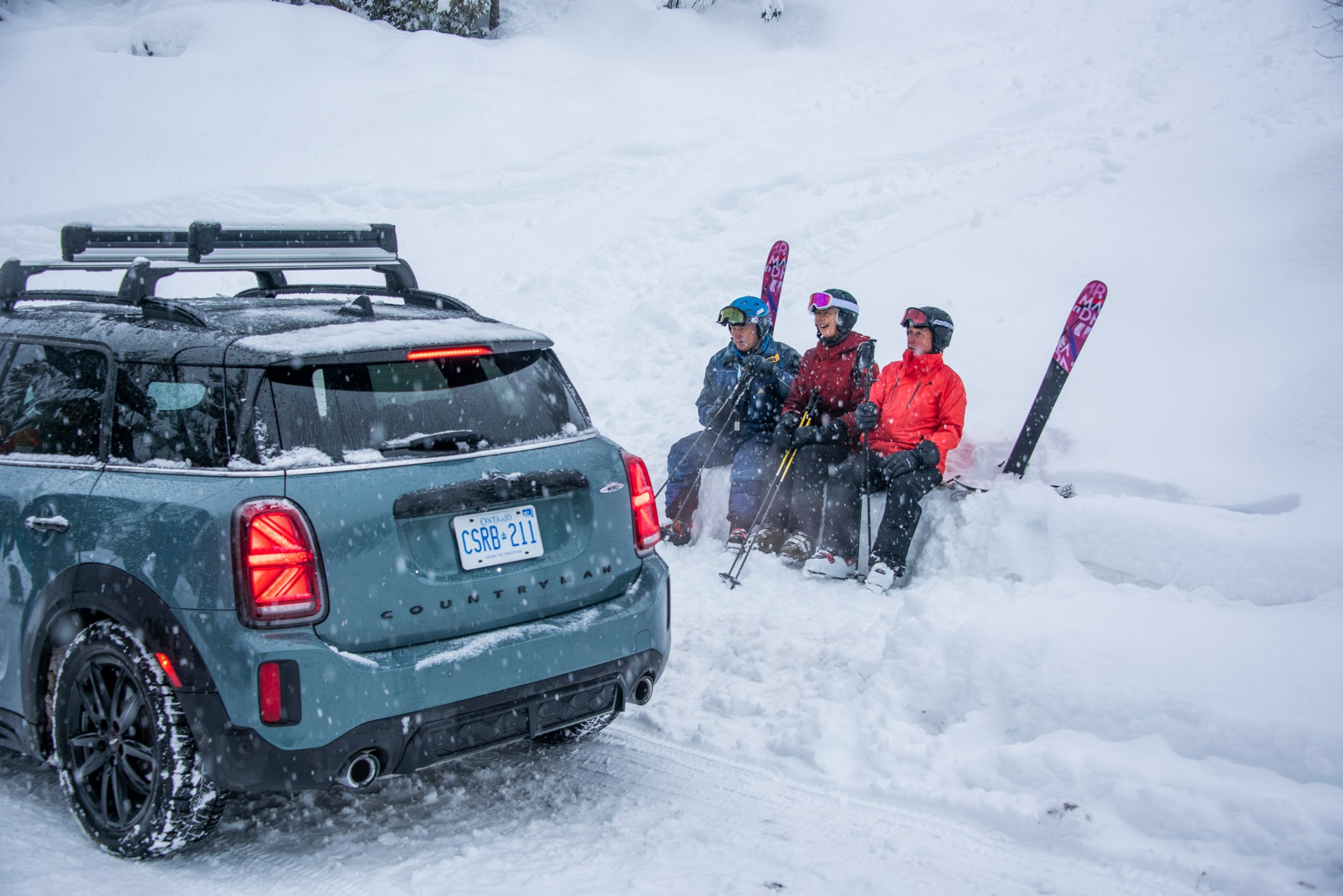 Image of three “Silver Sliders”, who are sitting in the snow with their ski equipment next to a MINI Countryman.
